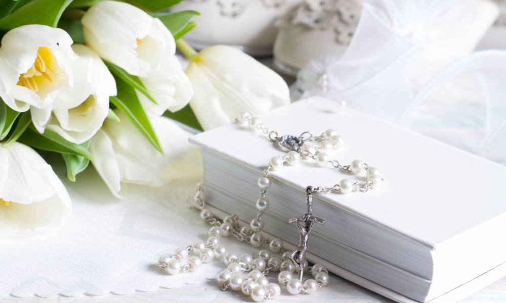 A white bible has a rosary sitting on top it and beautiful white flowers next to it. The rosary has iridescent pearl beads.