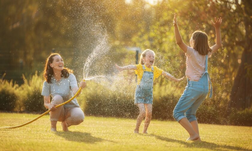 A mom outside with her two children playing in the yard with the water hose enjoying the summer sun.
