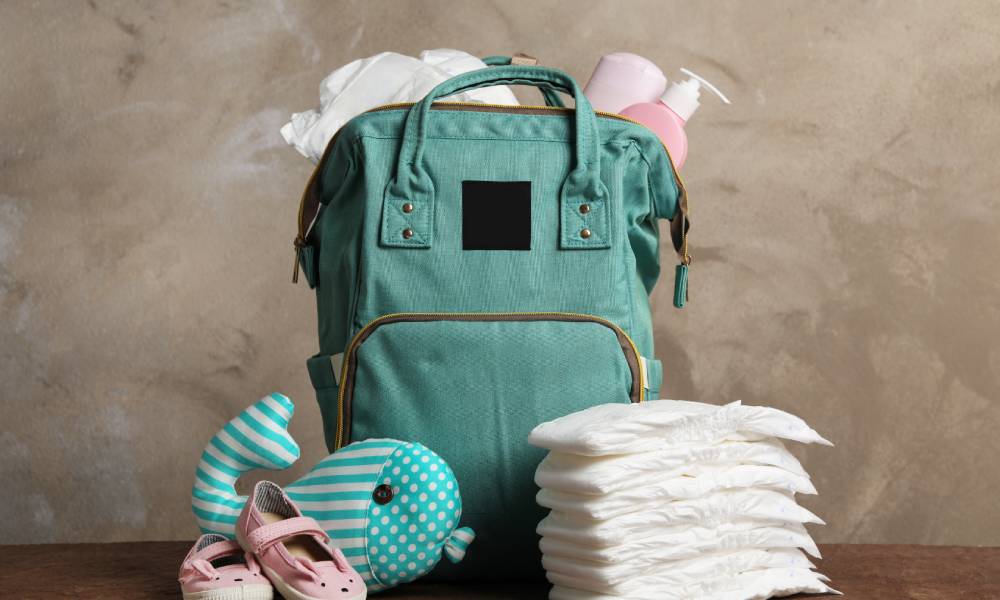 A diaper bag with lotion bottles and diapers peeking out the top. Extra diapers, baby shoes, and a stuffed toy sit at its base.