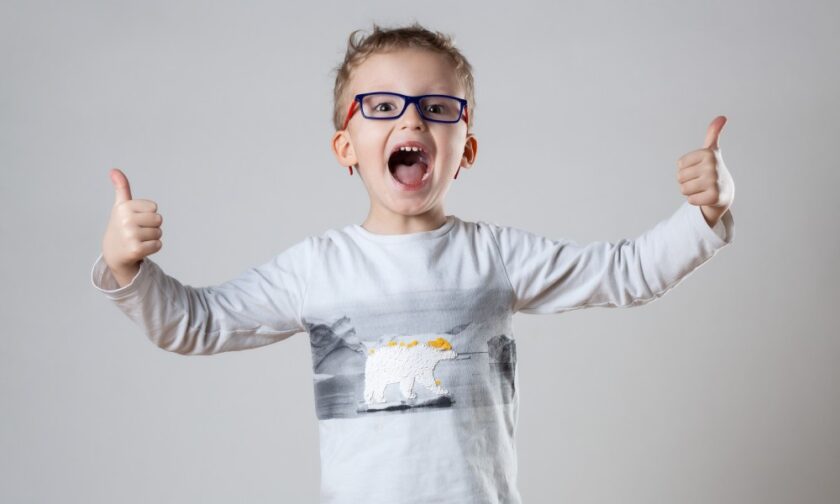 A young boy wearing a grey shirt gives the thumbs-up symbol with both hands. He's wearing blue and red glasses.