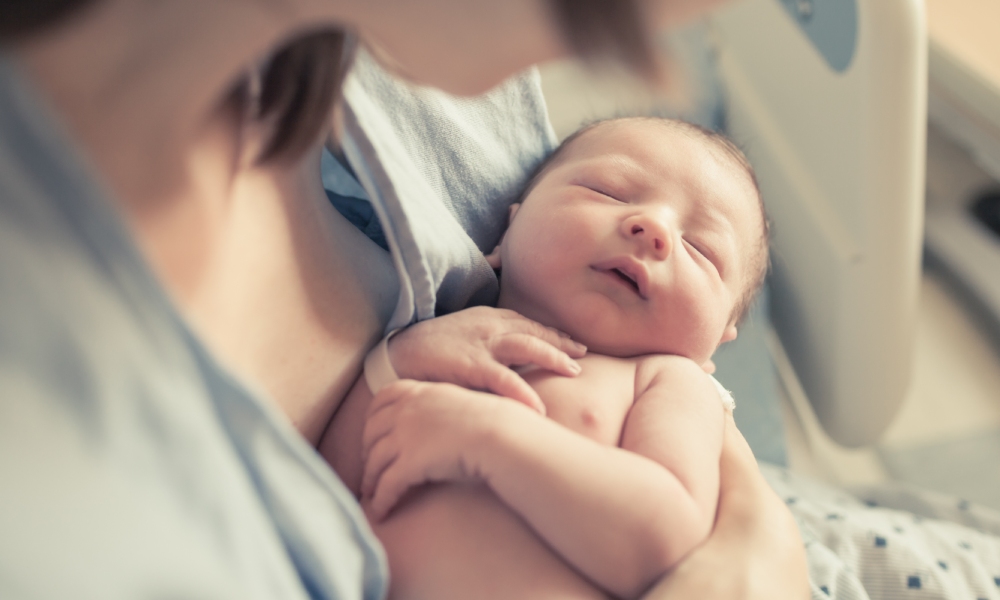 A mother holds her new baby in her arms while lying in the hospital bed. The new baby is asleep.