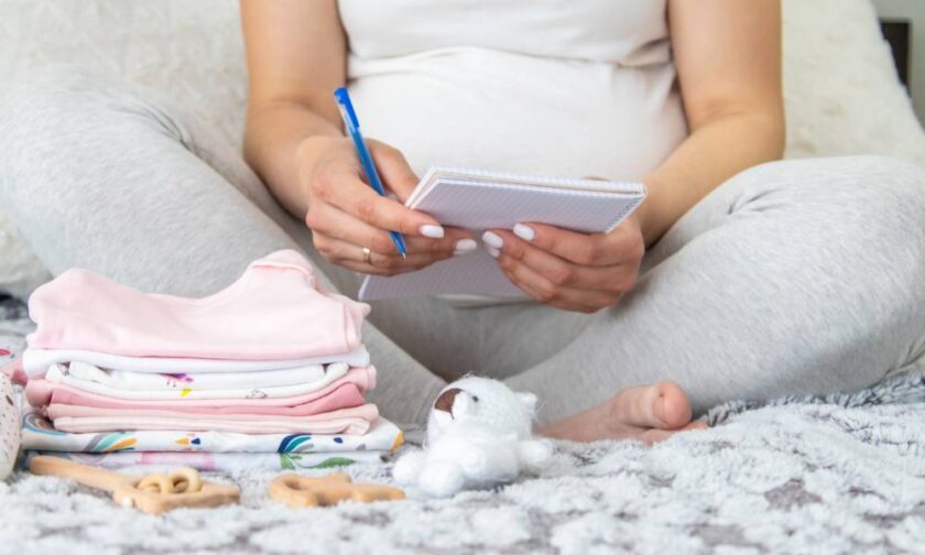 An expecting mother sitting down with a stack of baby clothes and a pen and notepad to create a birth plan.