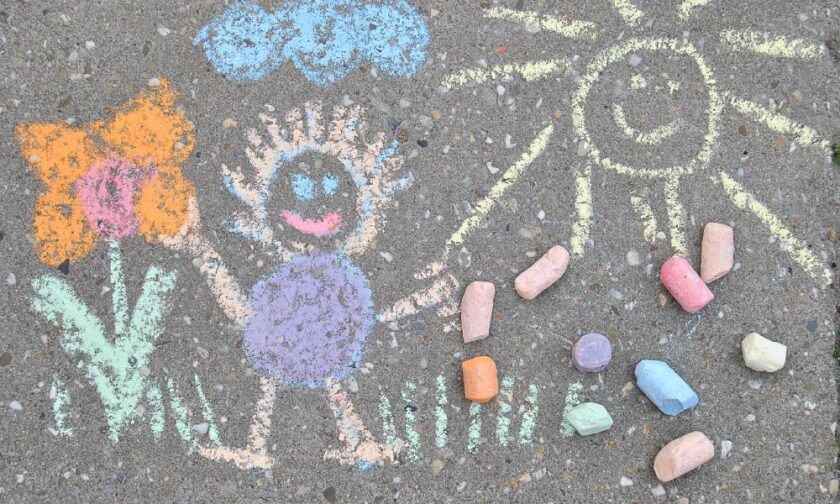 Sidewalk chalk drawings of a yellow sun and a little person with purple and orange limbs. There's also an orange flower.