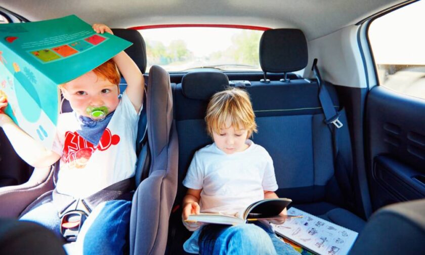 Two young children in the backseat of a car. One is in a child’s car seat, while the other is sitting and reading.