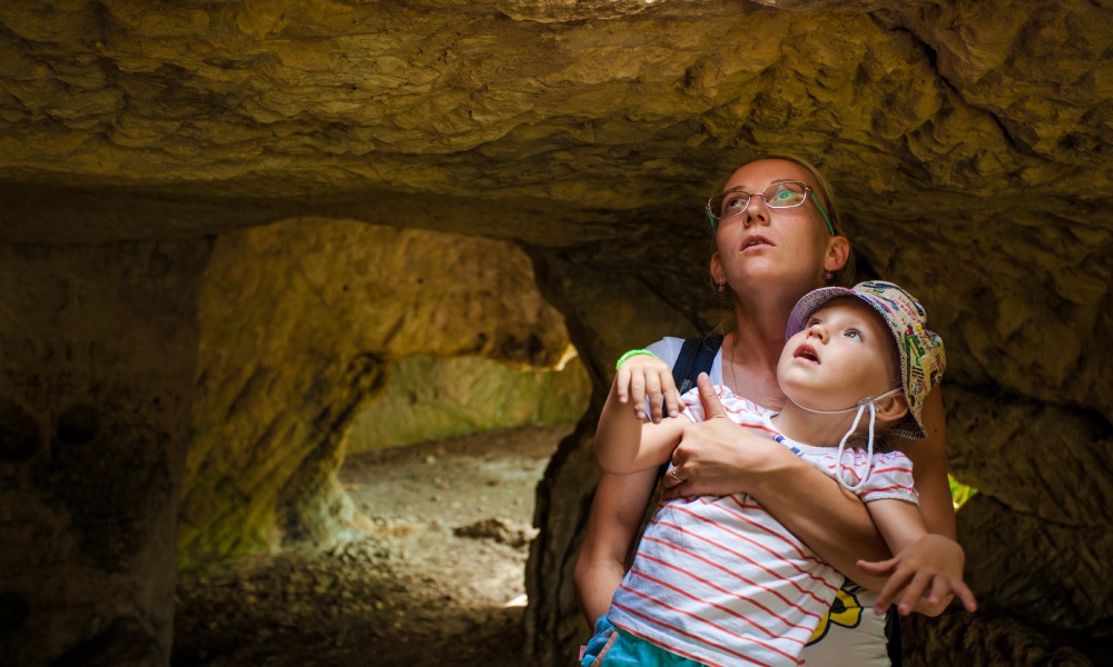 A mother hoisting up her child as they walk through a cave. Both stare up into a natural opening that lets light in.