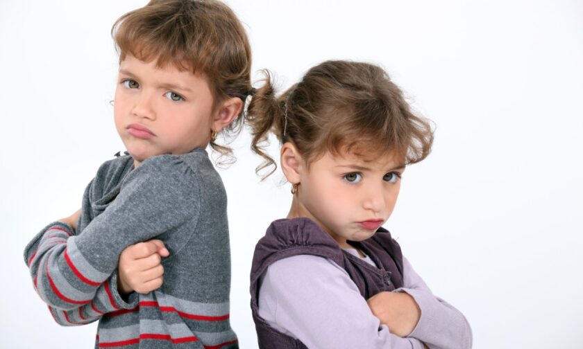 Two young girls with short pigtails standing back to back with their arms crossed. Both of them are pouting.