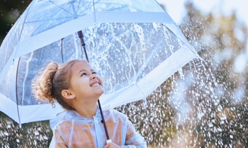 A young girl with a ponytail standing in the rain while holding a large clear umbrella and watching the droplets fall.