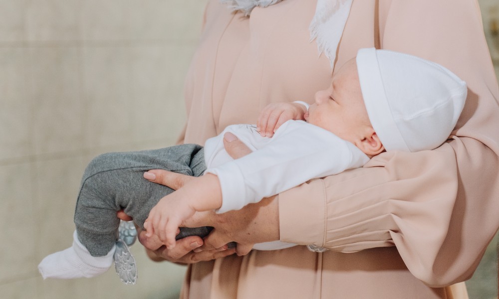 A close-up view of a godmother in a pink blouse holding a baby wearing neutral clothes and a beanie in her arms.