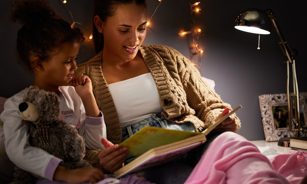 A mother sits with her child in bed, reading a story to the little girl. The daughter is in her pjs and holding a teddy bear.