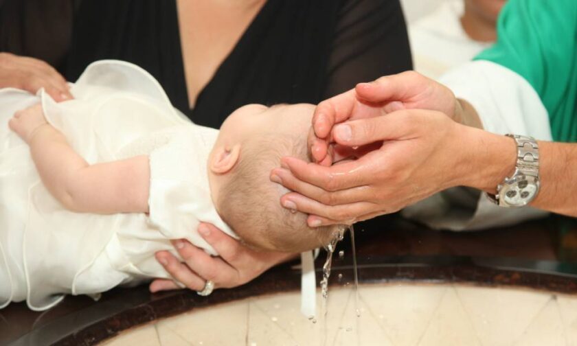 A close-up view shows a small baby in a white gown having water poured on its head. The baby's head is over a marble sink.