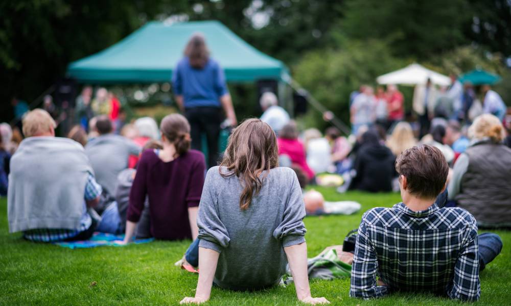 A crowd of people sit on a large patch of green grass. Green and white tents sit in front of the people.