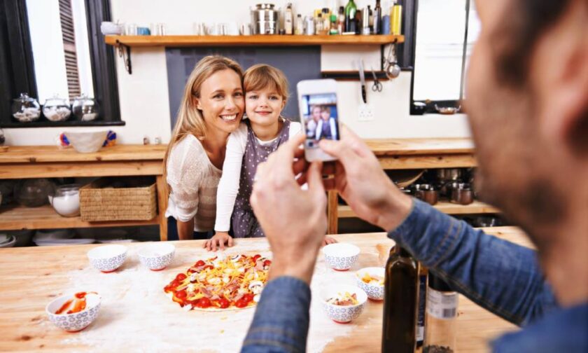 A father taking a photo of a mother and their daughter. The two are cooking together and smiling for a photo op.