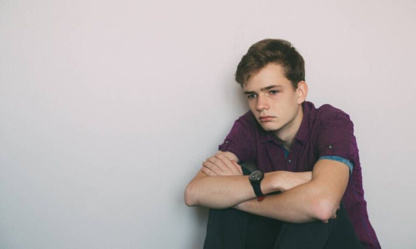 A young teen sitting against a blank wall and loosely holding his knees to his chest as he stares tiredly into the distance.