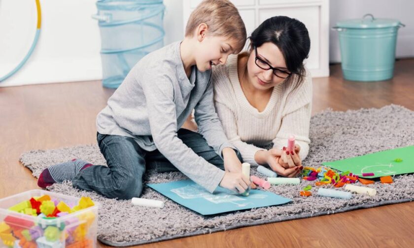 A young boy drawing using chalk on a blue sheet of paper. He and his mother lie on a gray rug on a wooden floor.