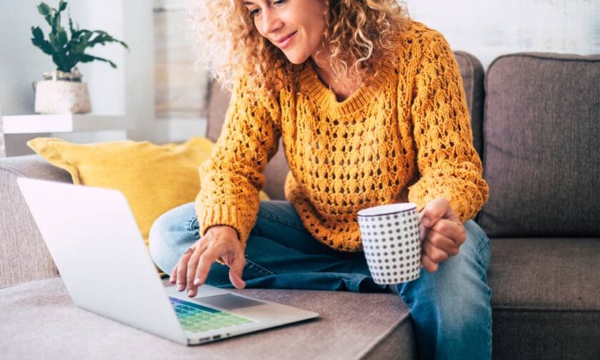 A woman in a yellow sweater sits on a couch. She has a mug in one hand and uses a laptop with the other.